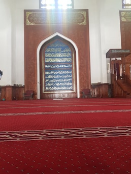 A large prayer hall with a richly decorated wooden mihrab featuring intricate calligraphy in gold on a blue background. The floor is covered in a red carpet with a dotted pattern, extending across the room. Two wooden chairs are positioned on either side of the mihrab, and a wooden staircase to the right appears to lead up to a raised platform.