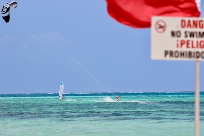 A windsurfer and a kite surfer are navigating through turquoise ocean waters. A red flag and a sign indicating 'danger' and 'no swimming' are prominently displayed in the foreground. The sky is clear and blue, complementing the sea.