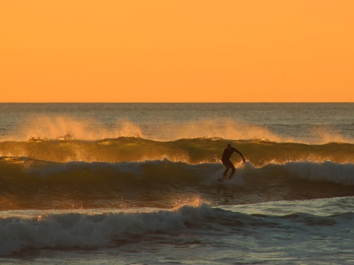 Sunset silhouette of a surfer catching a wave off Taghazout's rocky shore.