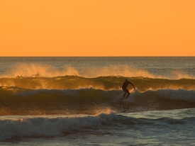A silhouette of a surfer riding a wave against a backdrop of a warm, orange sunset. The ocean spray is illuminated by the light, creating a glowing effect on the waves. The scene captures the dynamic movement of the surfer and the natural beauty of the seascape.