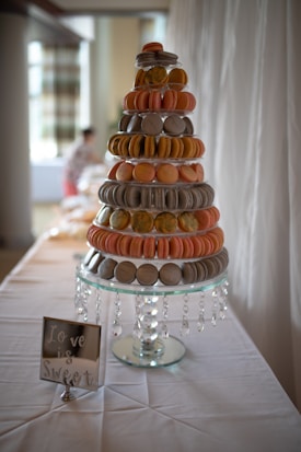 A multi-tiered macaron tower is displayed on a table. The tower consists of pink, orange, and gray macarons arranged neatly on a clear stand with hanging crystal-like decorations. Beside the tower, there is a small sign stating 'Love is Sweet'. The background is softly blurred, indicating a well-lit, possibly festive setting.