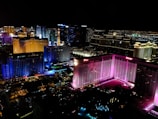 A vibrant cityscape at night featuring brightly lit buildings with diverse colors, including pink, blue, and gold. The skyline is marked by high-rise hotels and casinos, and the streets below are illuminated with lights.