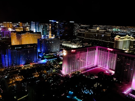 A vibrant cityscape at night featuring brightly lit buildings with diverse colors, including pink, blue, and gold. The skyline is marked by high-rise hotels and casinos, and the streets below are illuminated with lights.