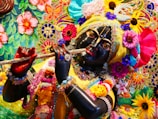 A detailed and colorful depiction of a divine figure adorned with vibrant floral garlands, elaborate embroidery, and rich jewelry, holding a flute. The figure's facial features are intricately crafted, with expressive eyes and a serene expression. The surrounding area is filled with a tapestry of colorful flowers and decorative patterns, contributing to a festive and spiritual atmosphere.