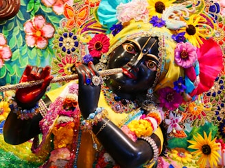 A detailed and colorful depiction of a divine figure adorned with vibrant floral garlands, elaborate embroidery, and rich jewelry, holding a flute. The figure's facial features are intricately crafted, with expressive eyes and a serene expression. The surrounding area is filled with a tapestry of colorful flowers and decorative patterns, contributing to a festive and spiritual atmosphere.