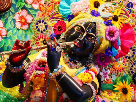 A detailed and colorful depiction of a divine figure adorned with vibrant floral garlands, elaborate embroidery, and rich jewelry, holding a flute. The figure's facial features are intricately crafted, with expressive eyes and a serene expression. The surrounding area is filled with a tapestry of colorful flowers and decorative patterns, contributing to a festive and spiritual atmosphere.