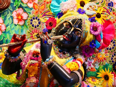 A detailed and colorful depiction of a divine figure adorned with vibrant floral garlands, elaborate embroidery, and rich jewelry, holding a flute. The figure's facial features are intricately crafted, with expressive eyes and a serene expression. The surrounding area is filled with a tapestry of colorful flowers and decorative patterns, contributing to a festive and spiritual atmosphere.