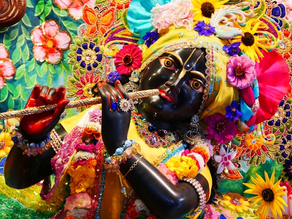 A detailed and colorful depiction of a divine figure adorned with vibrant floral garlands, elaborate embroidery, and rich jewelry, holding a flute. The figure's facial features are intricately crafted, with expressive eyes and a serene expression. The surrounding area is filled with a tapestry of colorful flowers and decorative patterns, contributing to a festive and spiritual atmosphere.