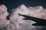An airplane wing slicing through thick, billowing clouds that are illuminated by soft, natural light. The scene captures the sense of flying high above the earth, surrounded by a vast expanse of white, fluffy clouds.