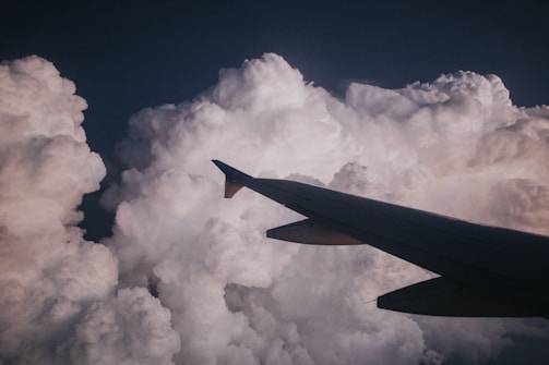 An airplane wing slicing through thick, billowing clouds that are illuminated by soft, natural light. The scene captures the sense of flying high above the earth, surrounded by a vast expanse of white, fluffy clouds.