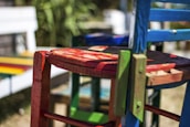 A close-up view of a collection of vibrantly painted wooden chairs. The focal chair is predominantly red with touches of green and blue visible in the frame. The chairs have an artistic, rustic charm and give off a handcrafted vibe. The background features a blurred scene with additional colorful elements, suggesting an outdoor setting.