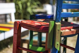 A close-up view of a collection of vibrantly painted wooden chairs. The focal chair is predominantly red with touches of green and blue visible in the frame. The chairs have an artistic, rustic charm and give off a handcrafted vibe. The background features a blurred scene with additional colorful elements, suggesting an outdoor setting.