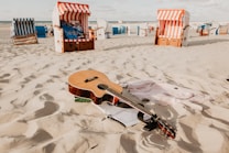 A sandy beach setting features a wooden acoustic guitar lying on the sand, surrounded by a few casual items, such as clothing. In the background, several colorful striped beach chairs, known as Strandkorb, are dispersed across the sand, suggesting a relaxed coastal atmosphere.