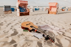 A sandy beach setting features a wooden acoustic guitar lying on the sand, surrounded by a few casual items, such as clothing. In the background, several colorful striped beach chairs, known as Strandkorb, are dispersed across the sand, suggesting a relaxed coastal atmosphere.
