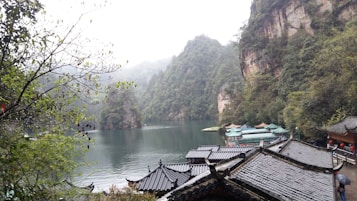 A serene landscape featuring a tranquil lake surrounded by lush green mountains. Traditional Chinese architecture with curved roofs is visible in the foreground, blending harmoniously with the natural surroundings. Mist lightly covers the mountain peaks, enhancing the peaceful and mystical quality of the scene.