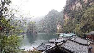 A serene landscape featuring a tranquil lake surrounded by lush green mountains. Traditional Chinese architecture with curved roofs is visible in the foreground, blending harmoniously with the natural surroundings. Mist lightly covers the mountain peaks, enhancing the peaceful and mystical quality of the scene.