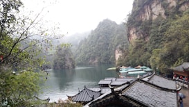 A serene landscape featuring a tranquil lake surrounded by lush green mountains. Traditional Chinese architecture with curved roofs is visible in the foreground, blending harmoniously with the natural surroundings. Mist lightly covers the mountain peaks, enhancing the peaceful and mystical quality of the scene.