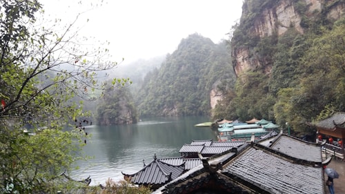 A serene landscape featuring a tranquil lake surrounded by lush green mountains. Traditional Chinese architecture with curved roofs is visible in the foreground, blending harmoniously with the natural surroundings. Mist lightly covers the mountain peaks, enhancing the peaceful and mystical quality of the scene.