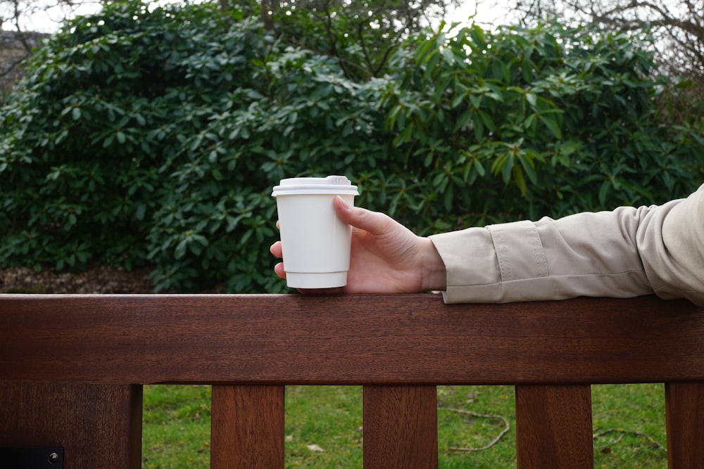 person holding white disposable cup on brown wooden railing