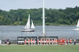 A group of athletes in orange and red jerseys is gathered on the sidelines of a sports field near a body of water. Sailboats are visible in the water, and there is a backdrop of lush green trees. The scene is sunny with a bright sky overhead.