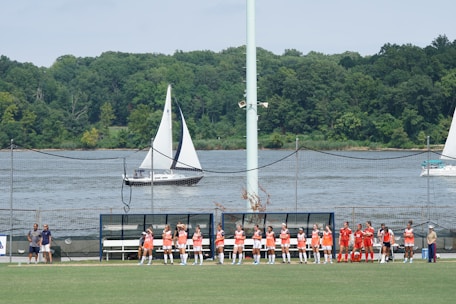 A group of athletes in orange and red jerseys is gathered on the sidelines of a sports field near a body of water. Sailboats are visible in the water, and there is a backdrop of lush green trees. The scene is sunny with a bright sky overhead.