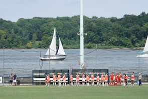 A group of athletes in orange and red jerseys is gathered on the sidelines of a sports field near a body of water. Sailboats are visible in the water, and there is a backdrop of lush green trees. The scene is sunny with a bright sky overhead.