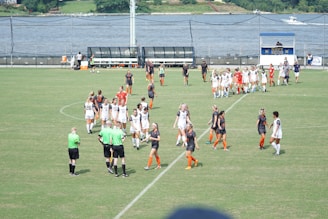 A women's soccer team in white uniforms and another in black and orange are gathered on a green soccer field. The players appear to be interacting with each other and with referees in green shirts. In the background, a body of water and a scoreboard are visible.