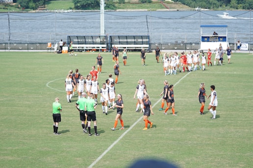 A women's soccer team in white uniforms and another in black and orange are gathered on a green soccer field. The players appear to be interacting with each other and with referees in green shirts. In the background, a body of water and a scoreboard are visible.
