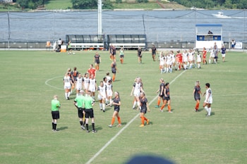 A women's soccer team in white uniforms and another in black and orange are gathered on a green soccer field. The players appear to be interacting with each other and with referees in green shirts. In the background, a body of water and a scoreboard are visible.