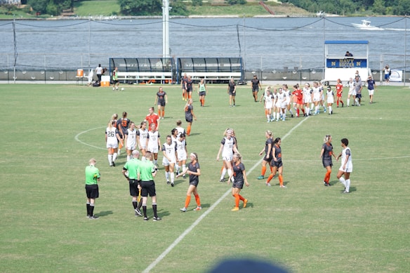 A women's soccer team in white uniforms and another in black and orange are gathered on a green soccer field. The players appear to be interacting with each other and with referees in green shirts. In the background, a body of water and a scoreboard are visible.
