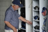 An elderly man with a white beard and wearing a navy cap and blue polo shirt is organizing or searching through a locker. The locker contains various headphones and personal items, some with patriotic designs featuring stars and stripes. The setting appears to be utilitarian, likely in a workplace or hobby area.
