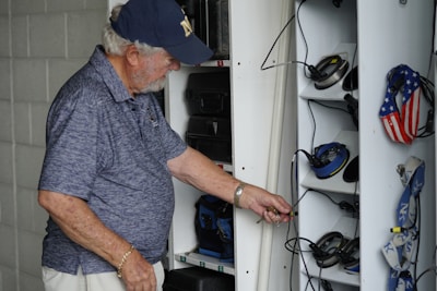 An elderly man with a white beard and wearing a navy cap and blue polo shirt is organizing or searching through a locker. The locker contains various headphones and personal items, some with patriotic designs featuring stars and stripes. The setting appears to be utilitarian, likely in a workplace or hobby area.