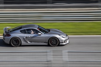 A sleek silver sports car is captured moving at high speed along a racetrack. The blurred background emphasizes the motion, with green grass and multiple horizontal barriers visible. The car features a spoiler and detailed design, showing precision and elegance.