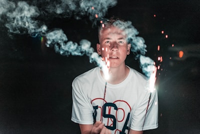 A person holding two sparklers in a dimly lit environment, surrounded by smoke from the sparklers. The individual is wearing a white t-shirt with the letters 'USA' on it.