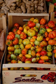 A wooden crate filled with a variety of colorful, small striped and multi-colored gourds and squash. Above the crate, there is another box containing ginger roots. The produce is displayed in a market setting.