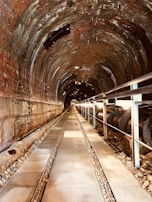A long, cylindrical tunnel is constructed with brick and metal materials. The tunnel is illuminated by natural light filtering through openings at the far end. There is a metal walkway with grated sections running along the center of the tunnel, with railings on the side. A large pipe runs parallel to the walkway, resting on gravel.