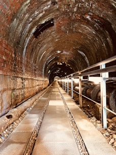A long, cylindrical tunnel is constructed with brick and metal materials. The tunnel is illuminated by natural light filtering through openings at the far end. There is a metal walkway with grated sections running along the center of the tunnel, with railings on the side. A large pipe runs parallel to the walkway, resting on gravel.