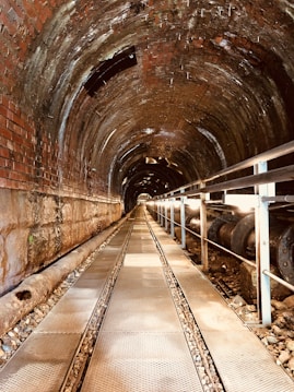 A long, cylindrical tunnel is constructed with brick and metal materials. The tunnel is illuminated by natural light filtering through openings at the far end. There is a metal walkway with grated sections running along the center of the tunnel, with railings on the side. A large pipe runs parallel to the walkway, resting on gravel.