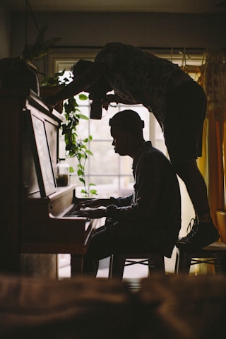 person sitting while playing piano while other man standing on stool while taking picture of person's hand on piano inside room