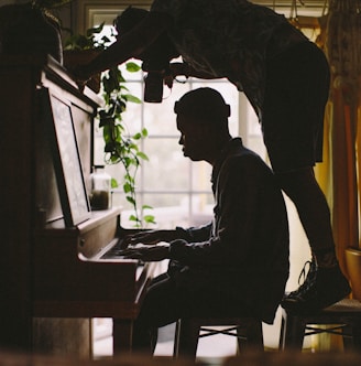 person sitting while playing piano while other man standing on stool while taking picture of person's hand on piano inside room