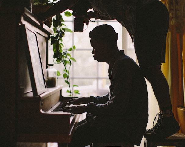 person sitting while playing piano while other man standing on stool while taking picture of person's hand on piano inside room