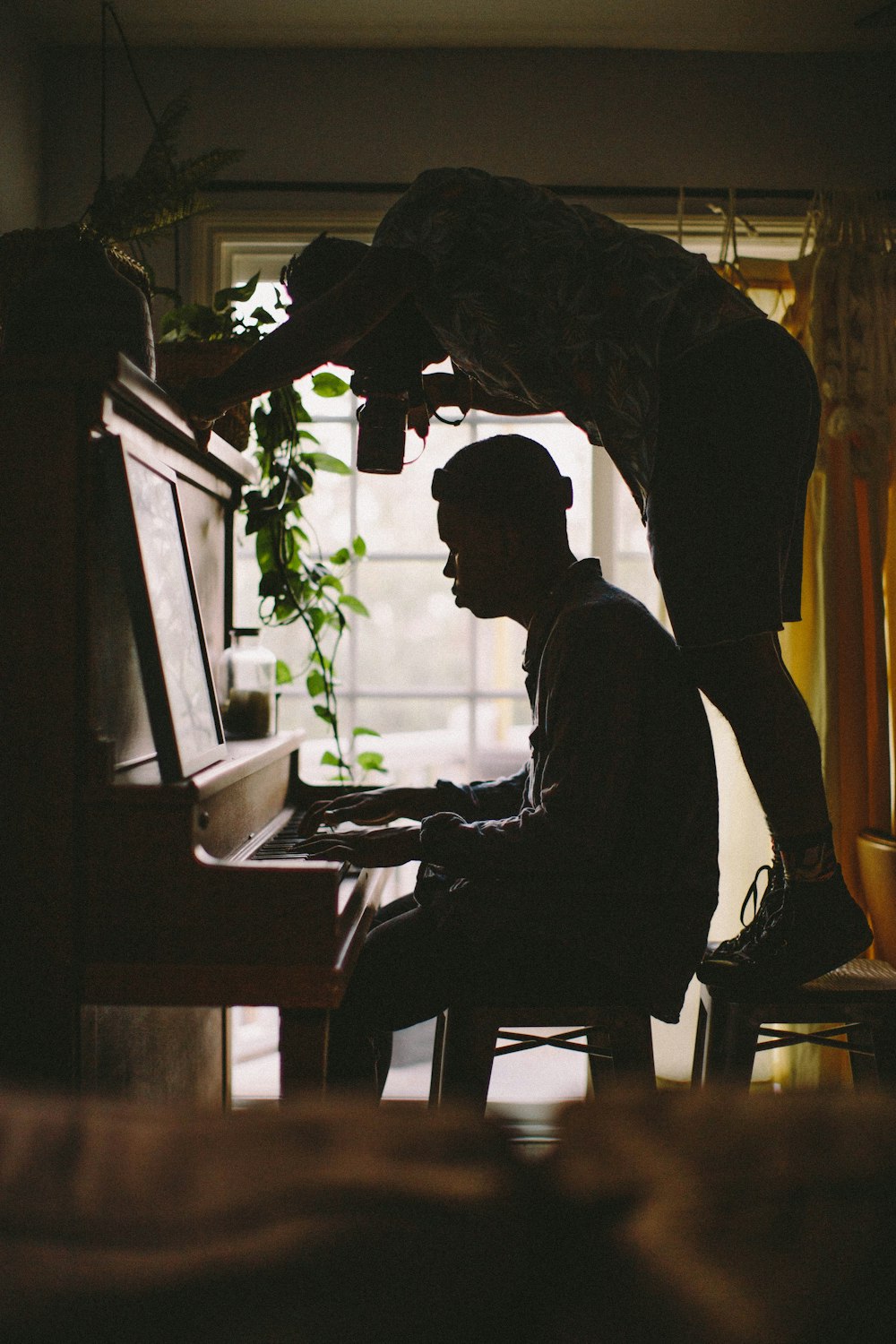 Person, die sitzt, während sie Klavier spielt, während ein anderer Mann auf einem Hocker steht, während sie die Hand einer Person auf dem Klavier im Zimmer fotografiert