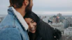 A couple smiling happily on a city rooftop with a panoramic view of a European skyline.