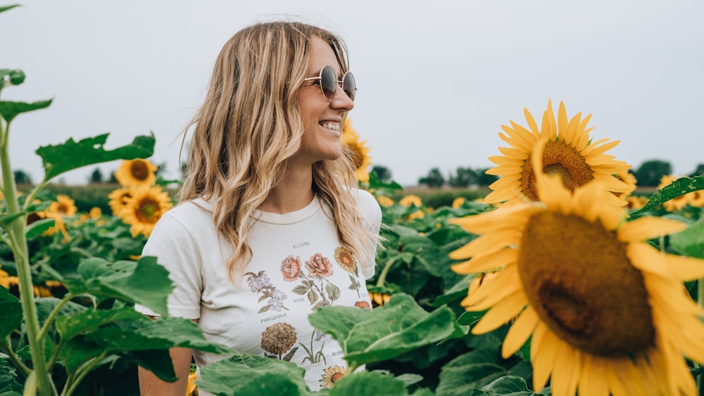 Frau in weißem und mehrfarbigem Blumenhemd steht und lächelt auf gelbem Sonnenblumenfeld