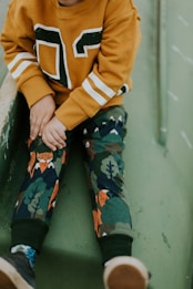 A child wearing a mustard-colored sweater with white and green accents, paired with pants featuring a playful forest and animal pattern. The setting suggests the child is sitting on a green slide or play structure. The hands are resting on the lap, and footwear with a mix of blue and brown tones is visible.