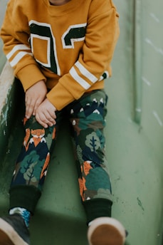 A child wearing a mustard-colored sweater with white and green accents, paired with pants featuring a playful forest and animal pattern. The setting suggests the child is sitting on a green slide or play structure. The hands are resting on the lap, and footwear with a mix of blue and brown tones is visible.