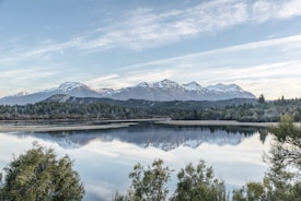A serene landscape with a calm lake reflecting the image of majestic snow-capped mountains under a light blue sky. Lush green trees frame the lake, and the scene conveys a sense of tranquility and natural beauty.