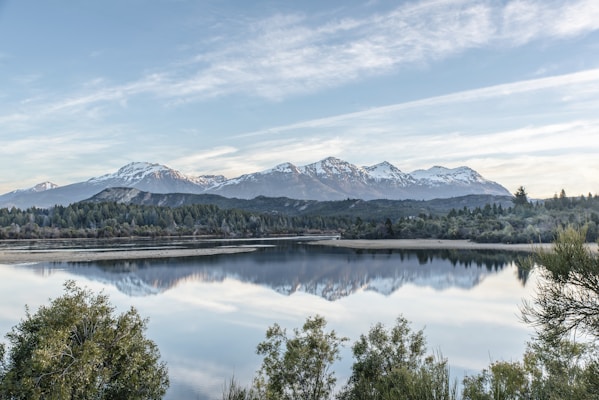 A serene landscape with a calm lake reflecting the image of majestic snow-capped mountains under a light blue sky. Lush green trees frame the lake, and the scene conveys a sense of tranquility and natural beauty.