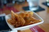 A white bowl is filled with crispy, golden-brown potato chips. The chips are arranged in an unorganized pile, creating a rustic and homemade appearance. In the background, there is a blurred view of a sleek smartphone and a glass of water on a wooden table with a red stripe running across it.