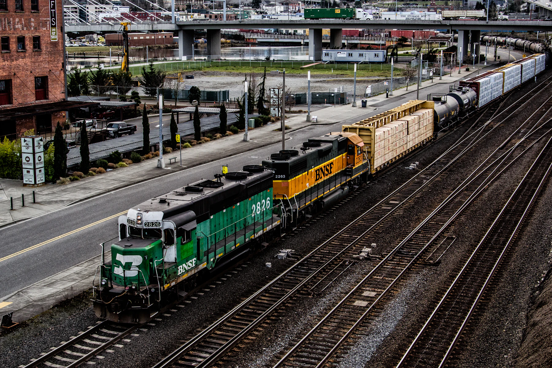A green and white diesel locomotive and an orange one pulling freight cars through a city rail corridor, seen from above